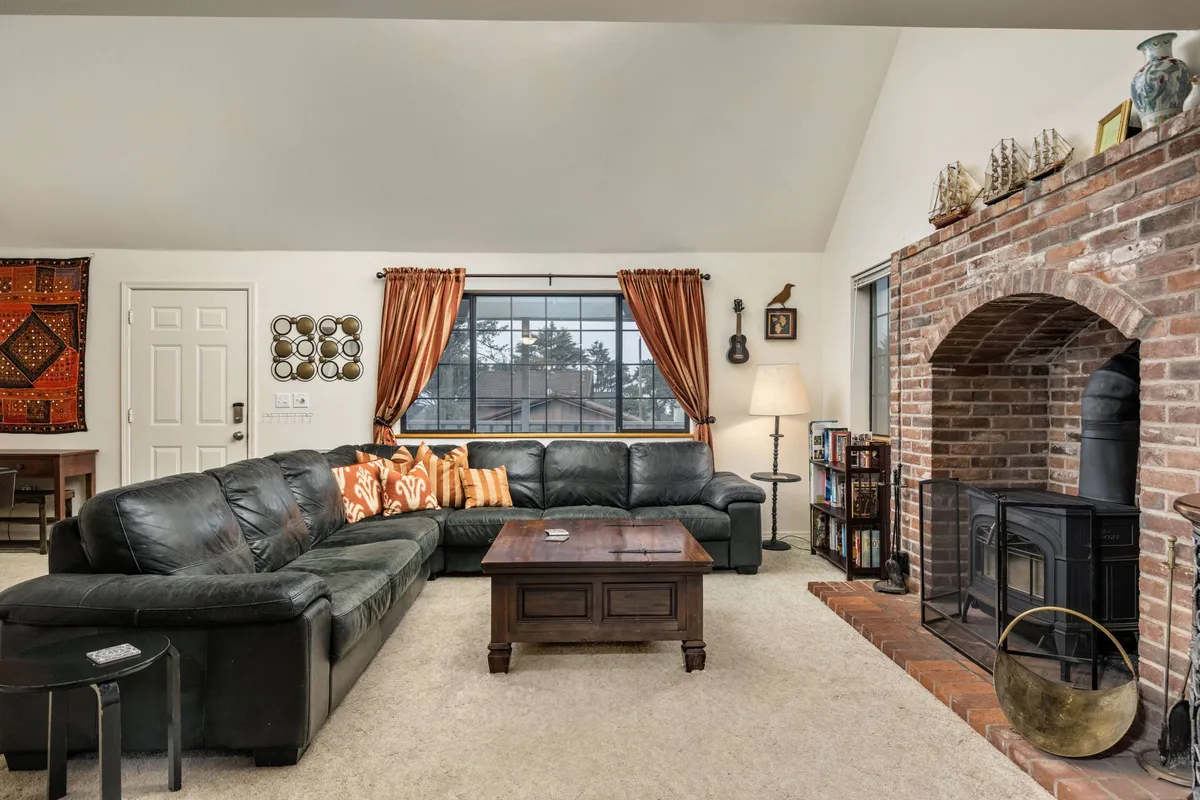 A spacious, sunlit living room featuring a large leather sectional sofa accented with colorful throw pillows, a brick fireplace that acts as a cozy focal point, and a wooden coffee table. Soft curtains frame the windows, allowing natural light to brighten the inviting space.
