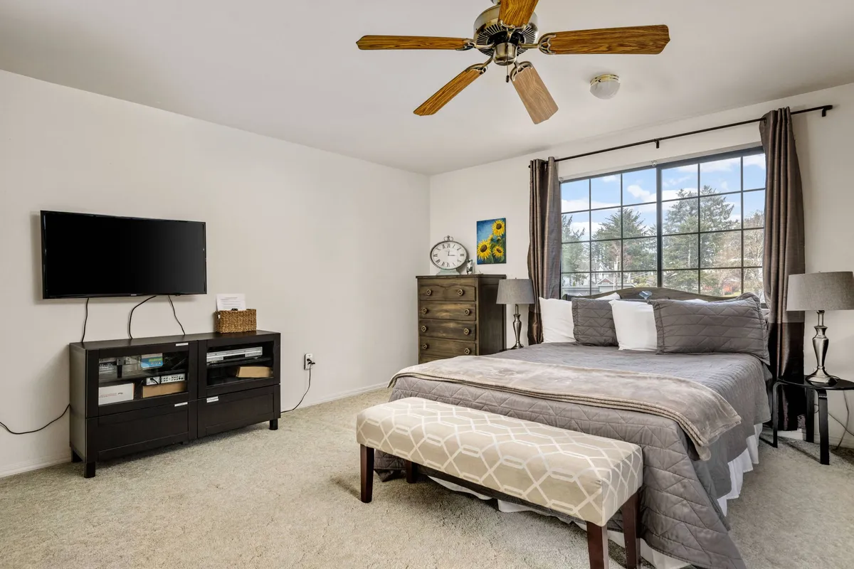 A calm upstairs bedroom with a bed dressed in gray bedding, a large window with curtains flooding the room with natural light, a wall-mounted TV for entertainment, a wooden dresser for storage, and a bench at the foot of the bed. The room feels both restful and practical for longer stays.