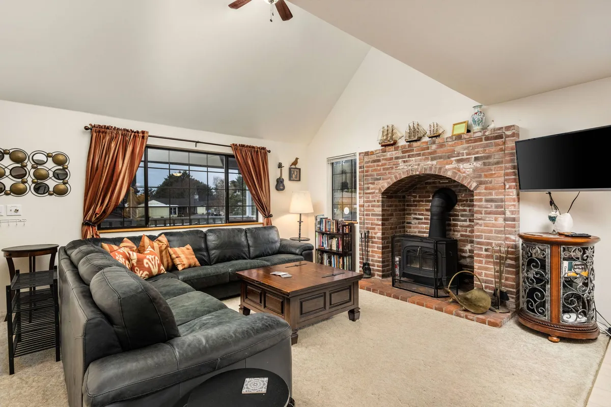 A comfortable living area featuring a large sectional sofa, a brick fireplace, and a flatscreen TV. Natural light streams in through curtained windows, highlighting the cozy and communal atmosphere of the space.