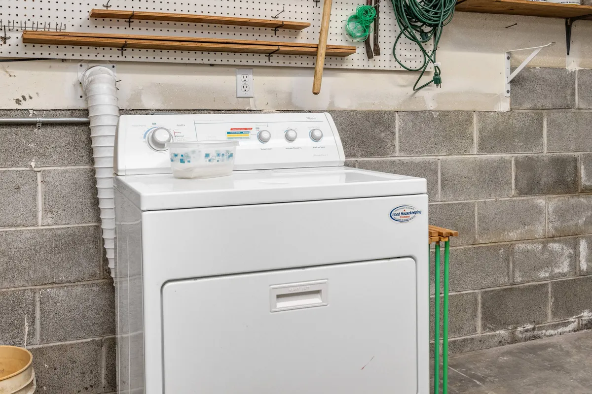 A close-up, practical view of a laundry area featuring a washing machine, shelving, and hanging cords. The utilitarian vibe emphasizes cleanliness and organization, showing the functionality of the shared laundry space.