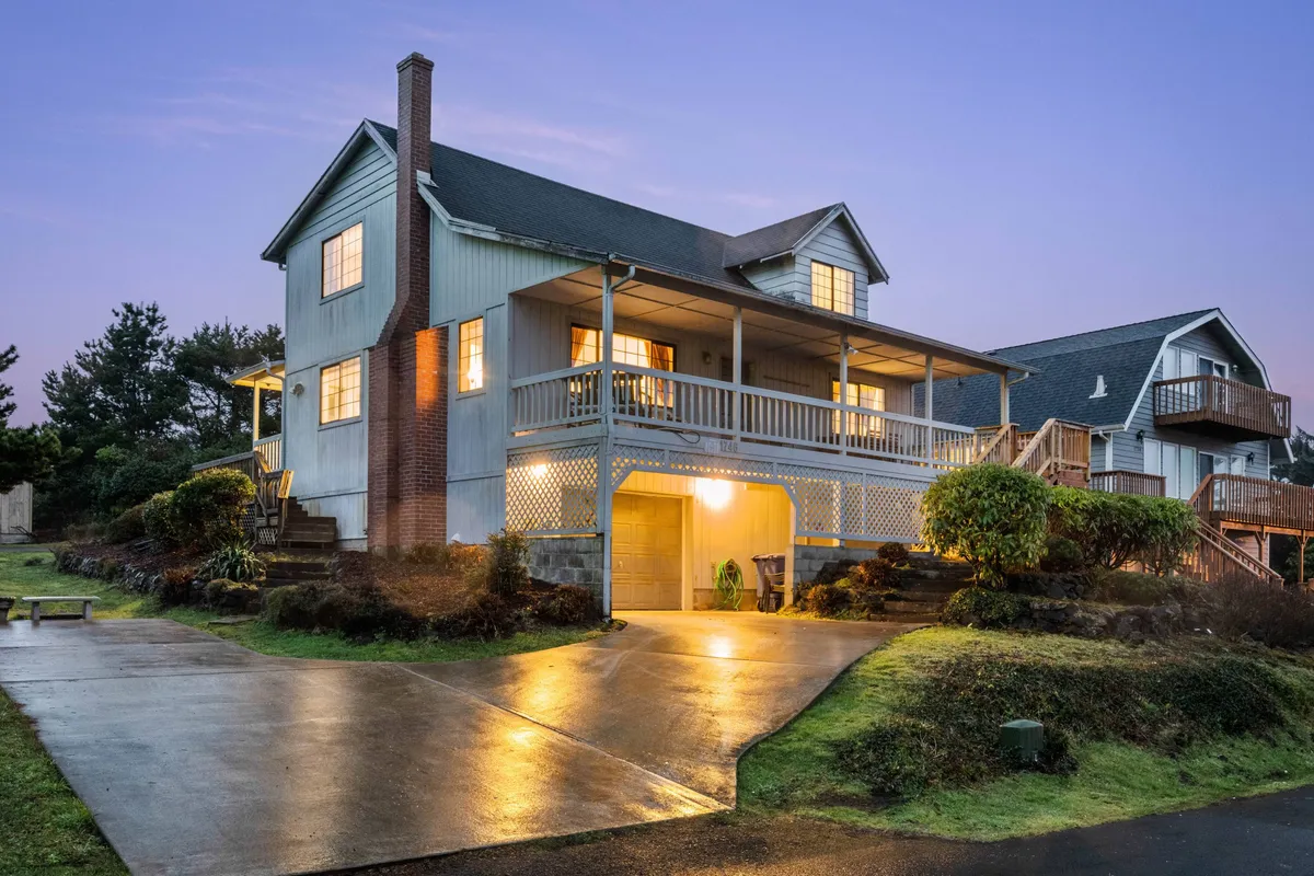 The image showcases the inviting exterior of a coastal single-family home in Lincoln City, Oregon. A spacious front porch with seating, large windows that let in natural light, and a generous driveway set a warm and welcoming scene. The porch is perfect for relaxing, entertaining, or soaking in the evening sunset.