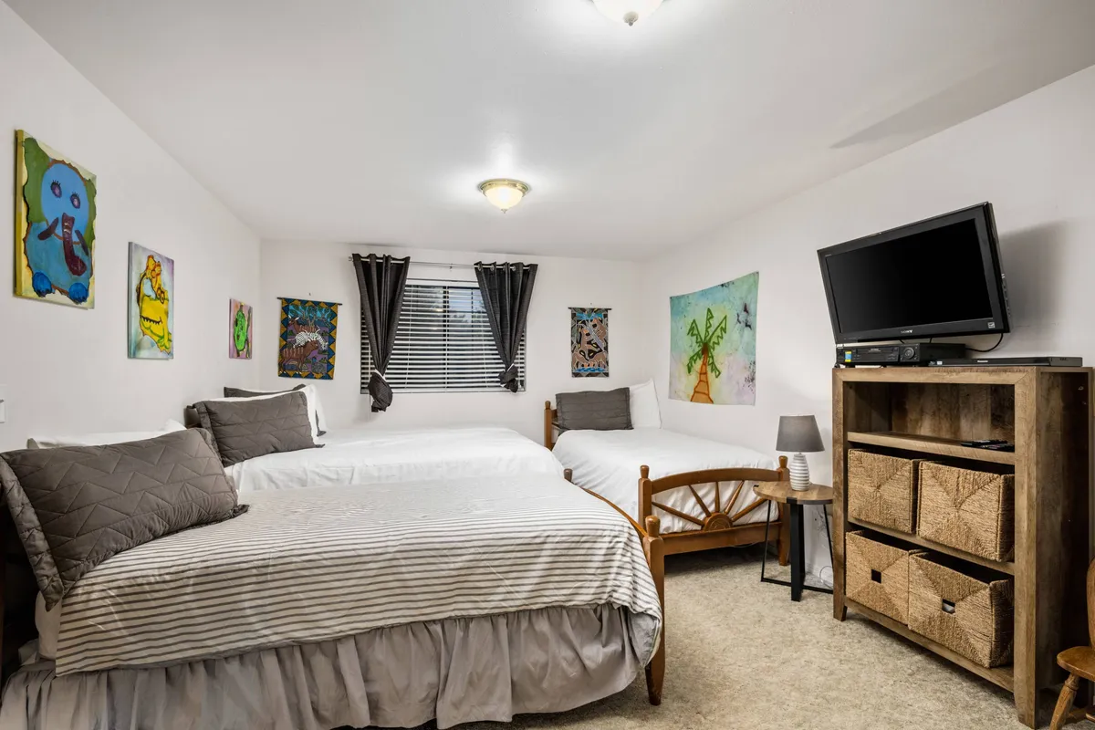 A playful, spacious bedroom showcasing three beds, colorful artwork, and wooden furniture with built-in storage. The setting is practical for families or groups, illustrating the home's flexible sleeping arrangements and creative ambiance, well-suited for children or groups.