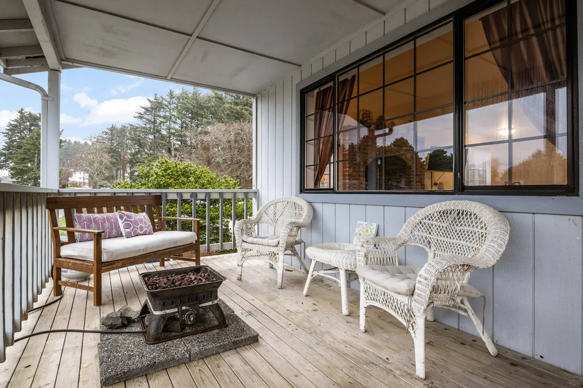 A cozy outdoor seating area featuring wicker chairs, a wooden bench, a fire pit, and a large window looking into the home. The space is surrounded by natural elements, offering a welcoming environment for relaxation, reading, or socializing.
