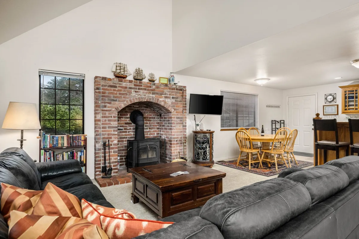 A cozy open-plan interior featuring a brick fireplace, a wooden coffee table, and an adjacent dining area with classic wooden chairs. The setup encourages both relaxation and socializing, perfectly blending comfort and utility.
